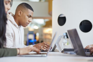 Three young adults are working together indoors, focused on laptops and collaborating on a project.