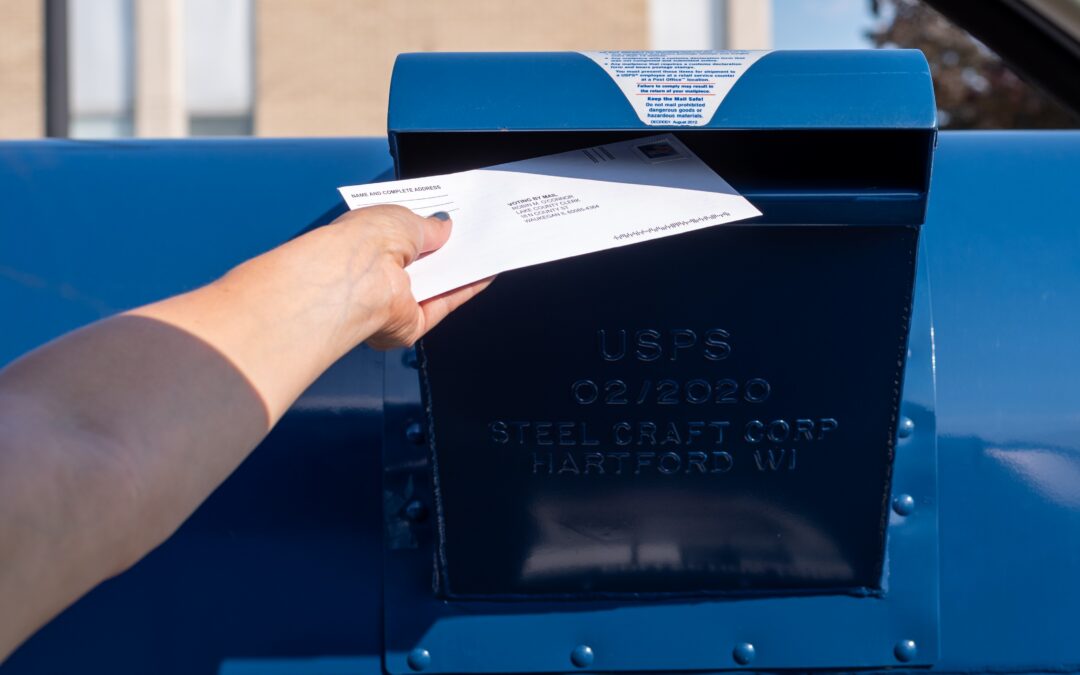 A hand places a white envelope into a blue United States Postal Service mailbox. The USPS emblem is visible on the front of the box, which is constructed of dark blue metal.