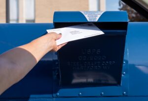 A hand places a white envelope into a blue United States Postal Service mailbox. The USPS emblem is visible on the front of the box, which is constructed of dark blue metal.