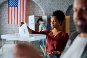 An adult woman drops a ballot into a transparent ballot box. The backdrop features an American flag and a brick wall with arched detailing.