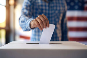 A hand in a blue patterned shirt places a ballot in a white box. The background is blurred with stripes of red and white. This image can be used for projects on the subject of voting, campaigns, or democracy. The lighting is bright and conveys a feeling of civic duty.